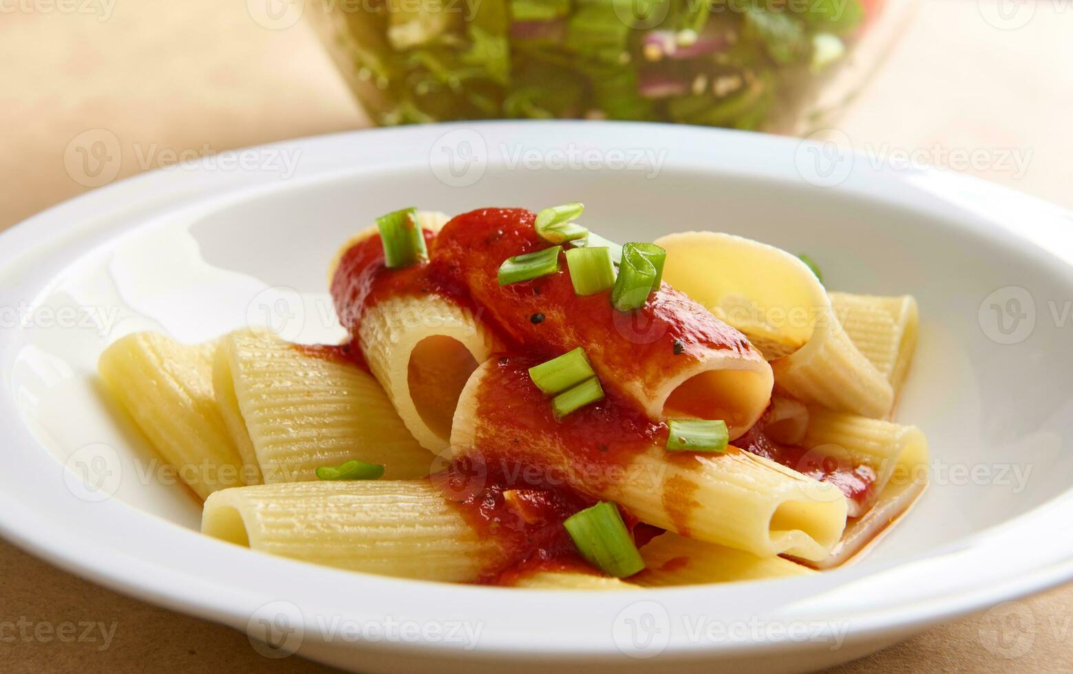 Healthy vegan meal of a traditional cooked Italian pasta with tomato sauce and green onion garnish against cropped blurred bowl with healthy herb salad photo