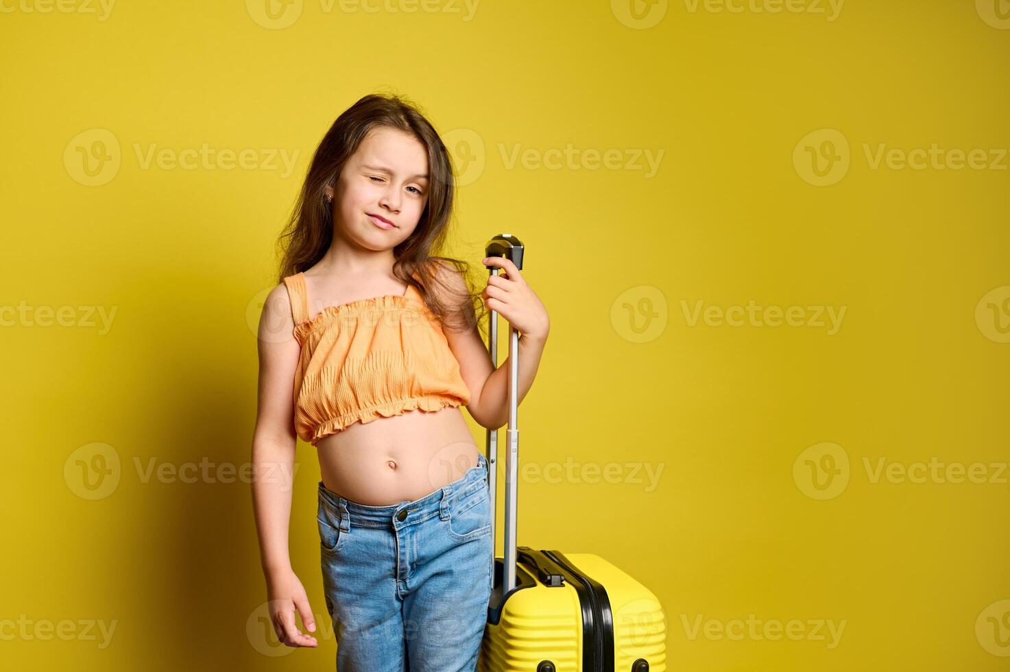 Cute girl winking at camera, traveling abroad, going for weekend getaway, posing with yellow suitcase. Studio background photo