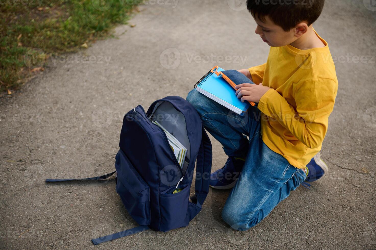 Charming elementary aged schoolboy sits on his knees on a path in the park, puts a notebook and pencil case in a backpack, returning home after shooting photo