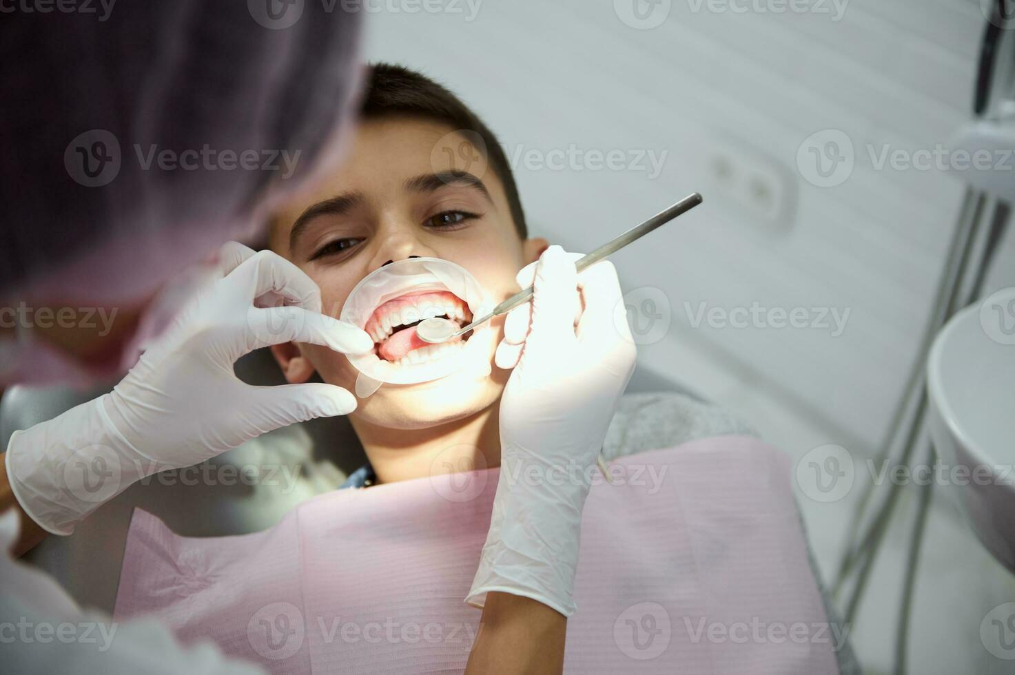 parte superior vista. de cerca de pequeño chico vistiendo un dental mejilla retractor durante regular médico chequeo. dentista utilizando inoxidable acero dental espejo y examinando pequeño chico dientes a dental cita foto