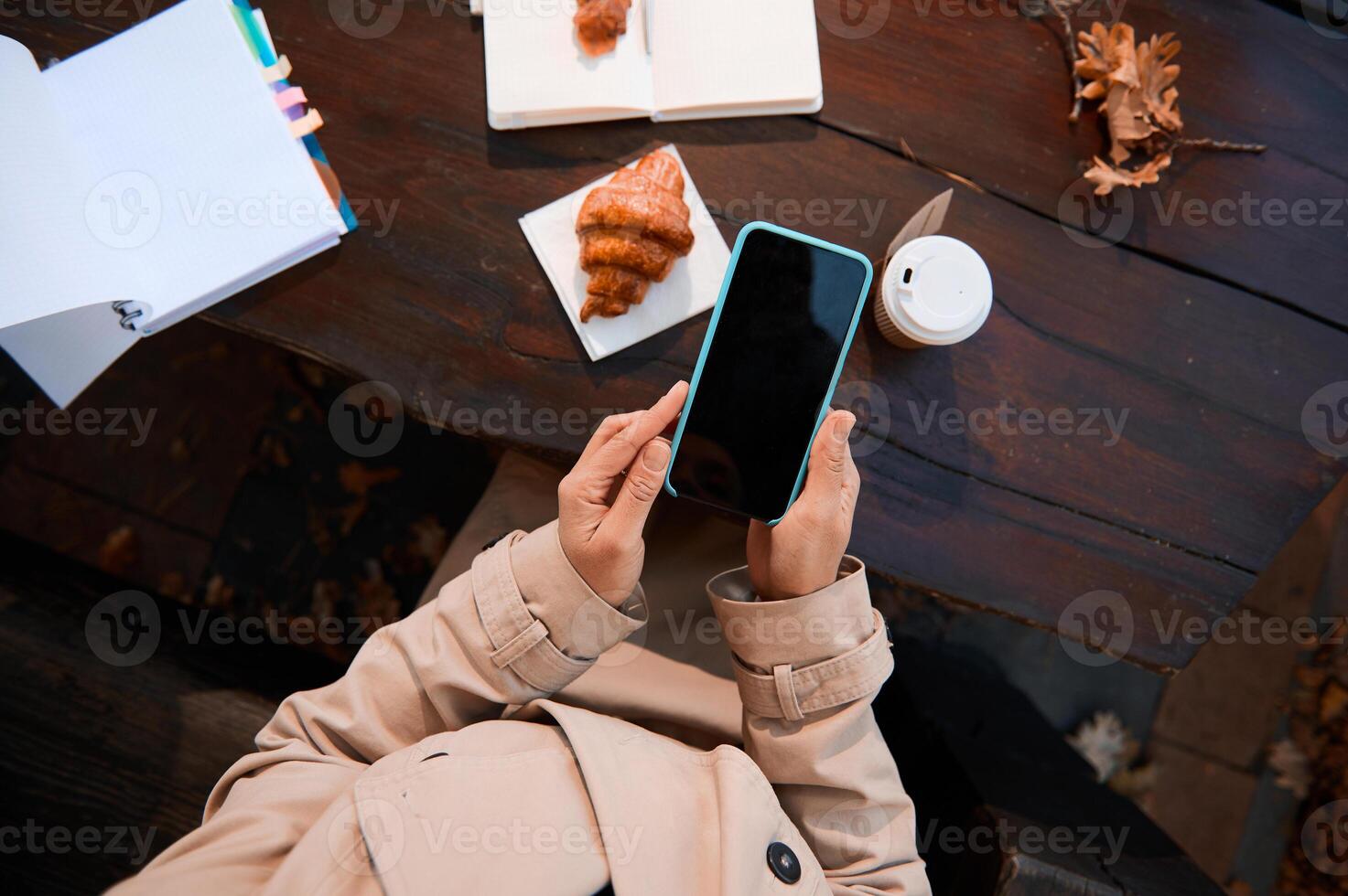 Close-up of an unrecognizable person hands using a mobile phone with blank black screen for copy space on the background of a wooden table with open notepad and copybook and delicious breakfast photo