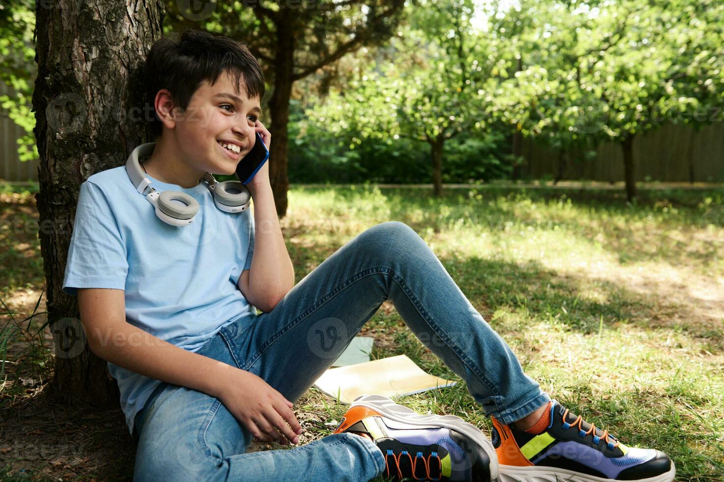 Cheerful handsome teen boy talking on mobile phone, wearing headphones, smiling, relaxing sitting under tree in the park photo