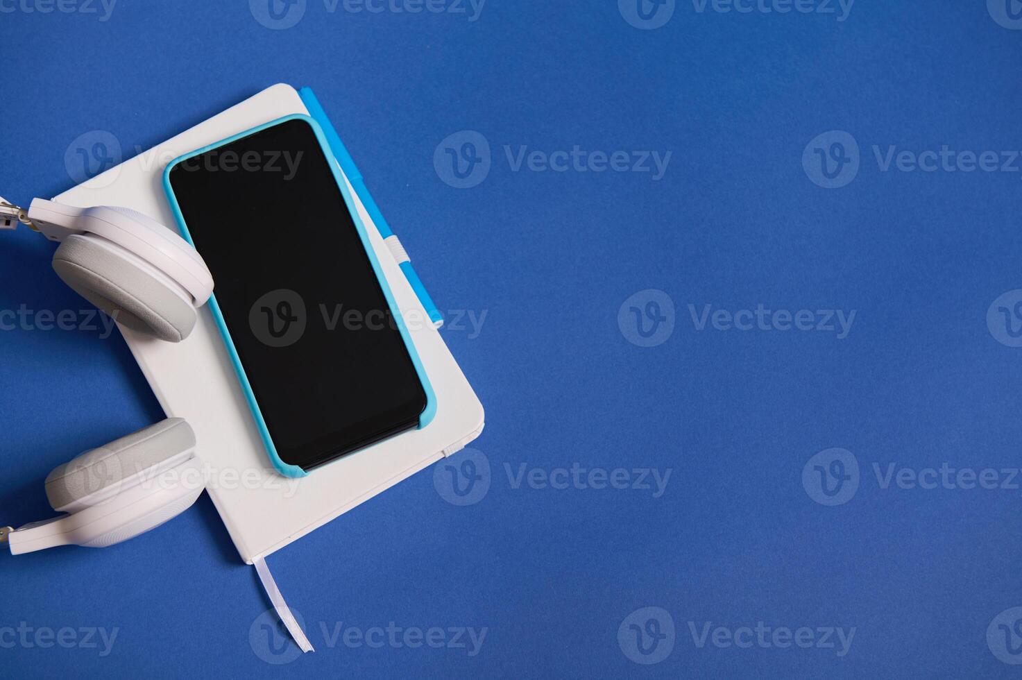 Flat lay of headphones, smartphone in blue case on white diary organizer isolated on blue background with copy space. Top view of white and blue office tools photo