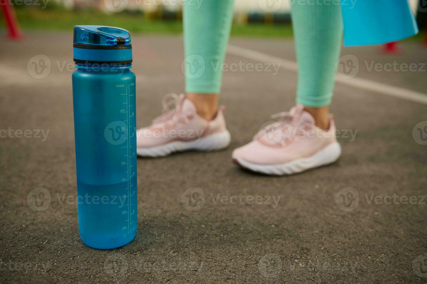 Close-up of a blue plastic bottle of water on the asphalt of sports ground next to sporty woman's feet. Health and body care concept photo