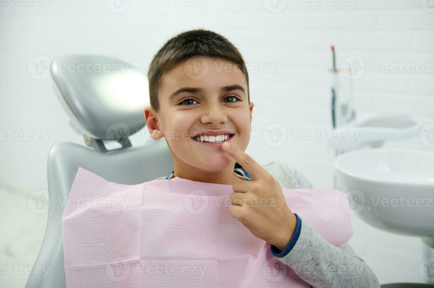 Cheerful boy holds finger near his mouth, looks at camera, smiles with beautiful toothy smile after receiving dental treatment in dentistry clinic. Oral hygiene, early prevention teeth disease concept photo