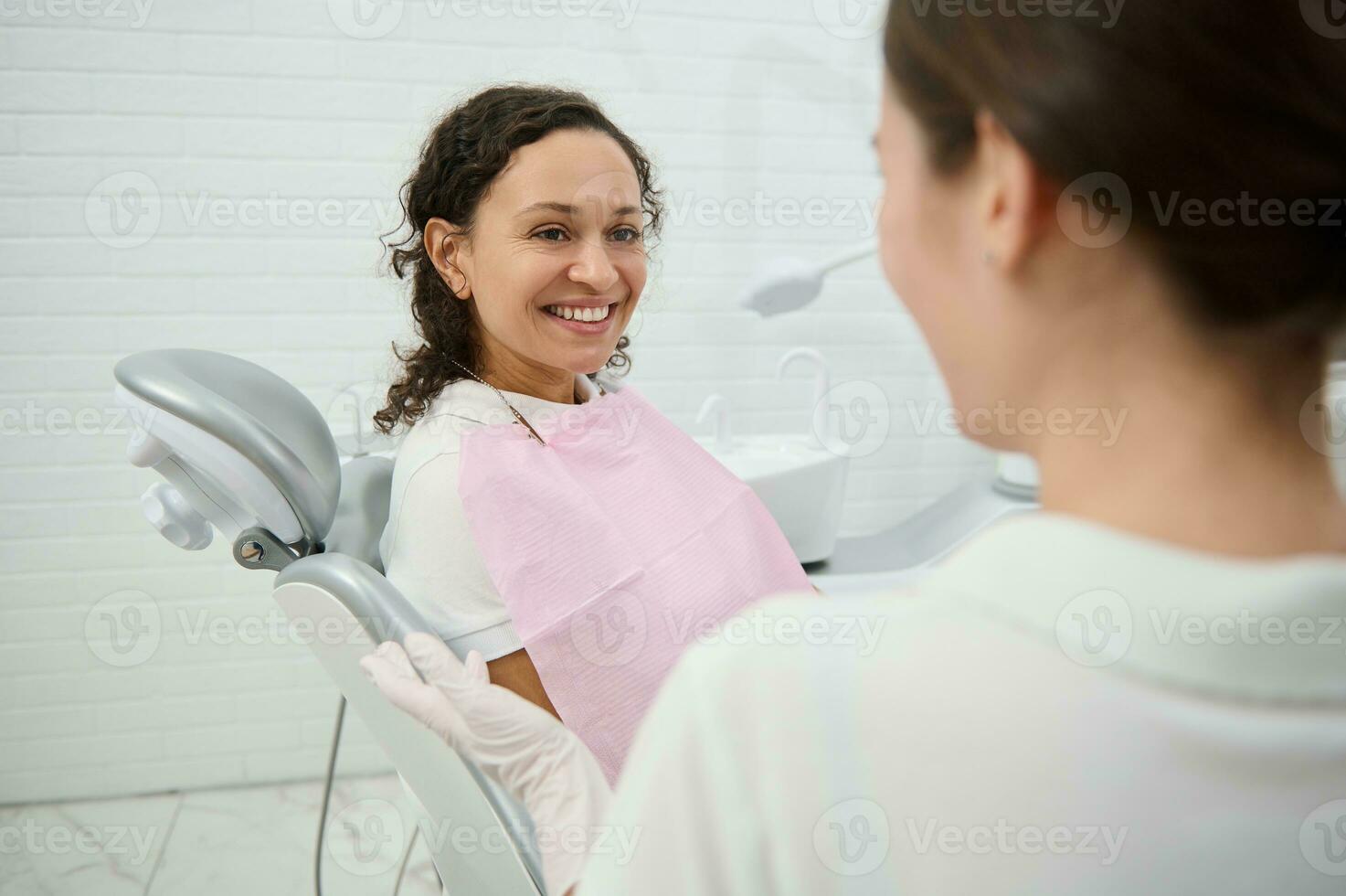 Cheerful African American woman sitting in dentists chair smiles toothy smile looking at dental assistant preparing her for regular medical check-up during regular dental appointment photo