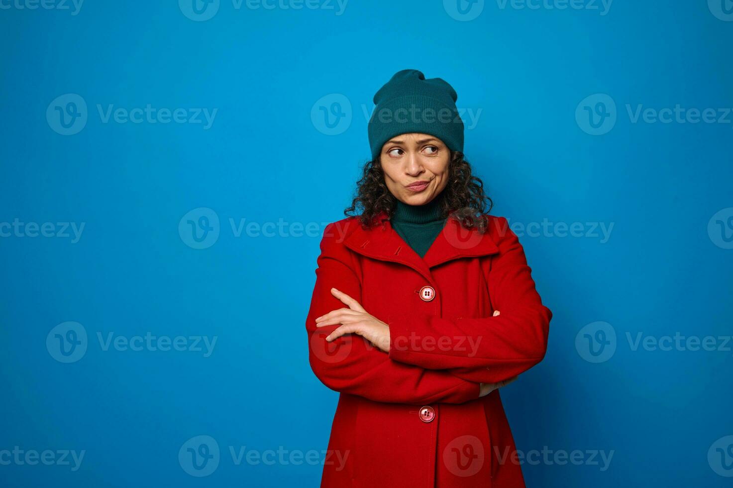 Portrait of a puzzled thoughtful pensive curly dark-haired pretty African American woman in red coat and green looking aside posing with crossed arms against blue wall background with copy ad space photo