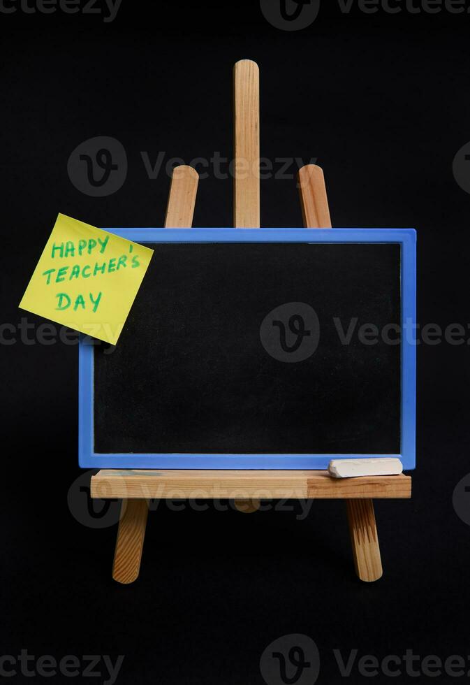 Close-up of a wooden table easel with chalk and blackboard with empty blank space for text and pasted note paper with inscription teachers day isolated on black background with copy space photo
