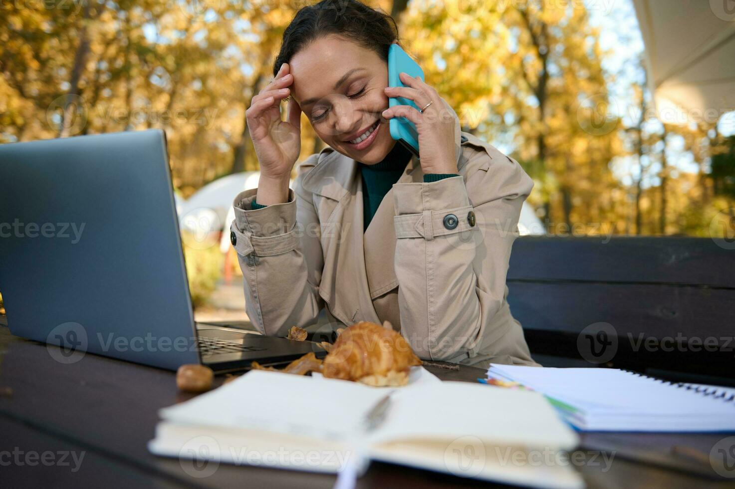 Attractive dark-haired business woman in a beige trench coat smiles toothy smile talking on mobile phone enjoying remote work away from hustle and bustle of the city on autumn oak grove background photo