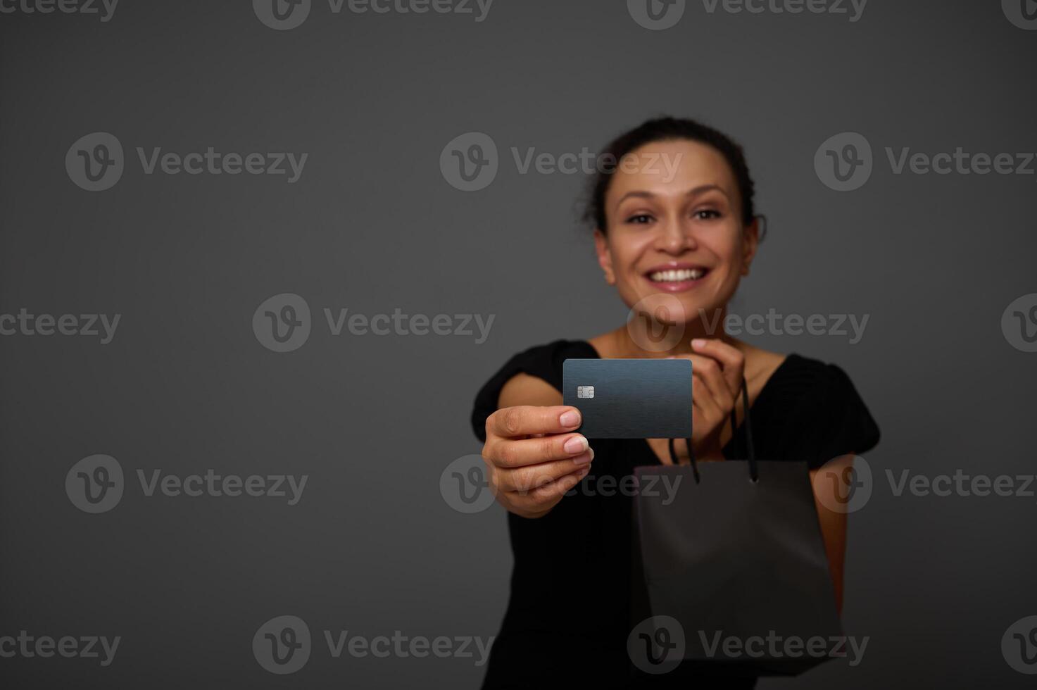 Soft focus on black credit card in the hand of smiling joyful woman dressed in black and posing with black shopping packet against gray wall background with copy space for Black Friday ad photo