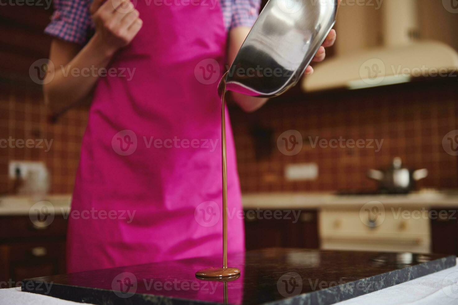 Confectioner chocolatier in pink apron pouring chocolate mass from bowl into marble surface for tempering. photo