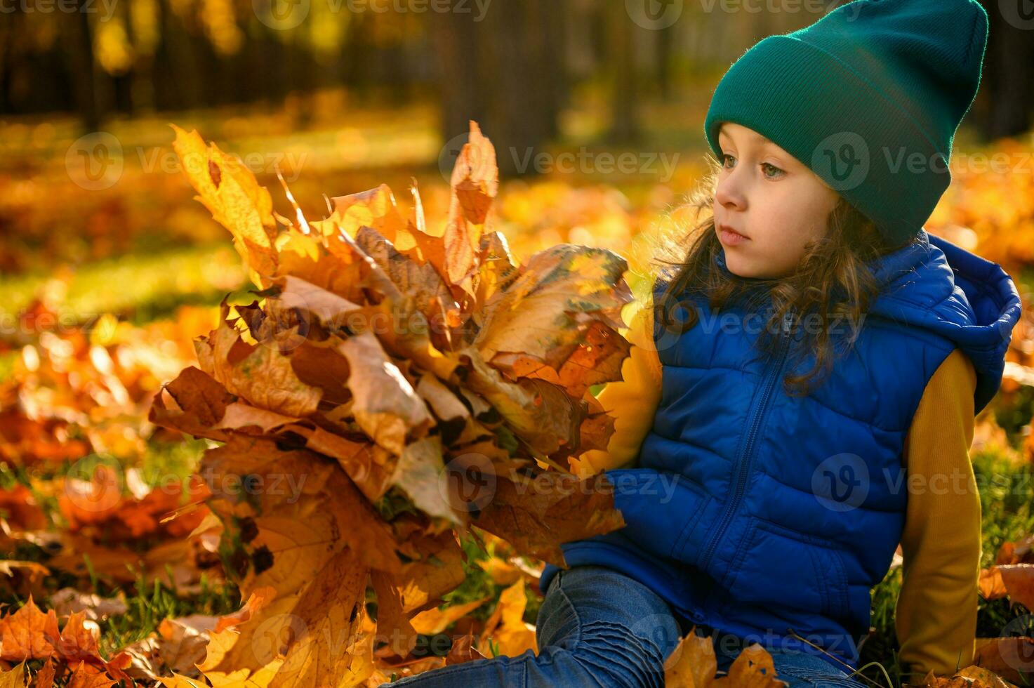 Pensive beautiful baby girl in bright colorful clothes posing with yellow bouquet of dry maple leaves sitting among fallen leaves against autumnal nature background with copy space for advertisement photo