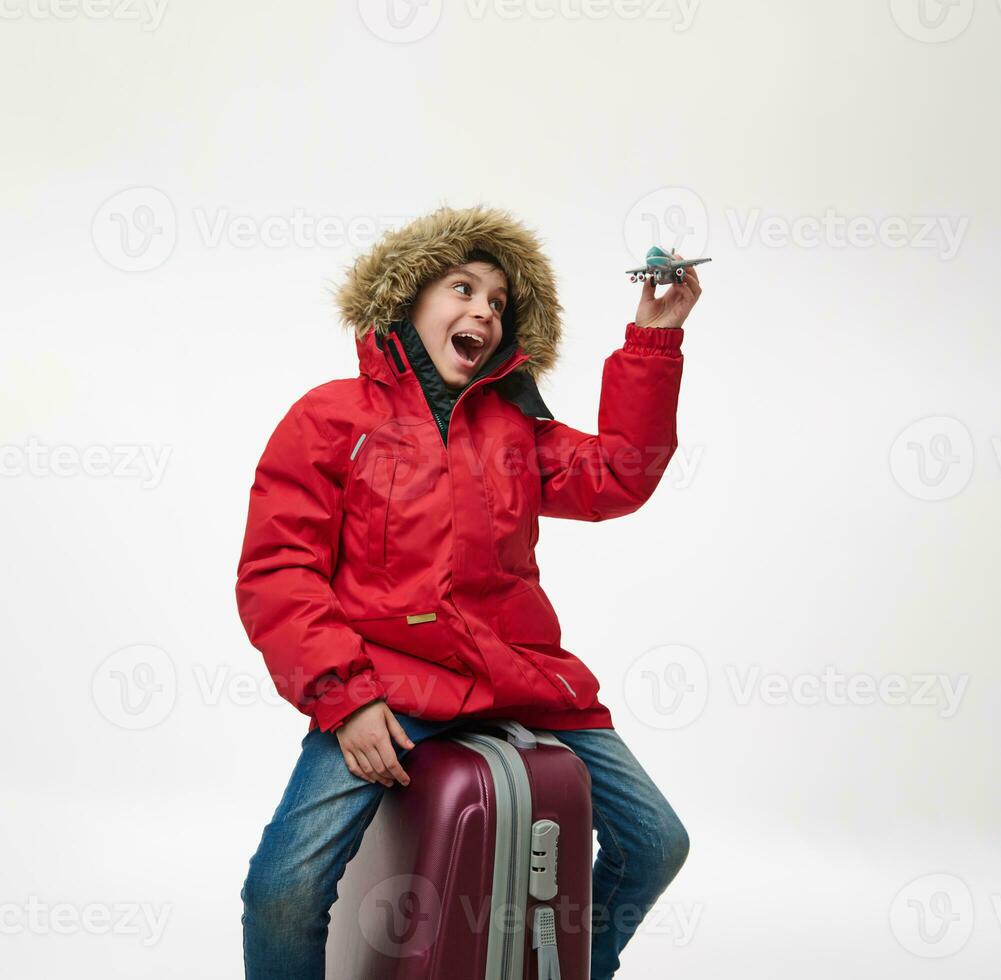 Adorable school boy in warm red down jacket with hood, sitting on a suitcase and playing with a toy model of an airplane, imitating the flight in the sky. Isolated on white background, copy ad space photo