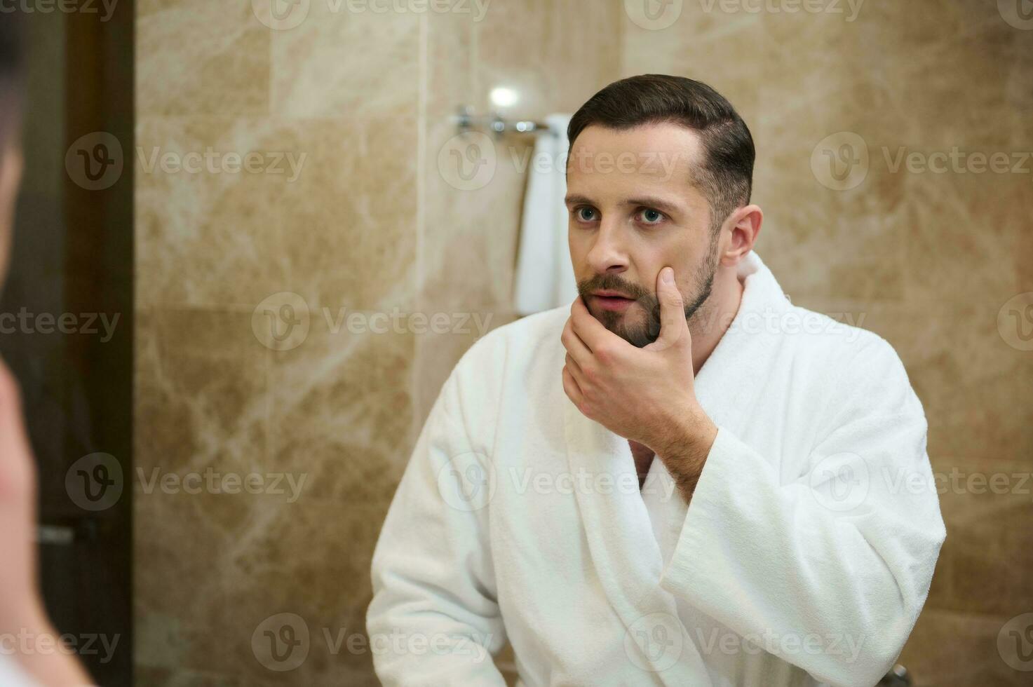Handsome young European man in a white terry bathrobe touching the stubble before shaving, looking in surprise at his mirror reflection in the bathroom photo
