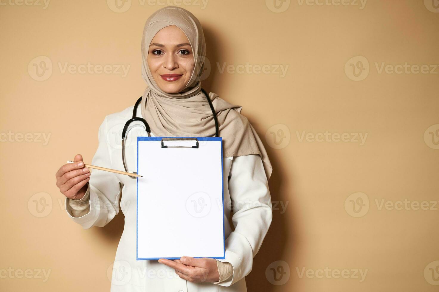 Charming female doctor in hijab pointing with a pencil at a blank sheet of paper on a clipboard. Healthcare and insurance concept. Copy space for promotion photo