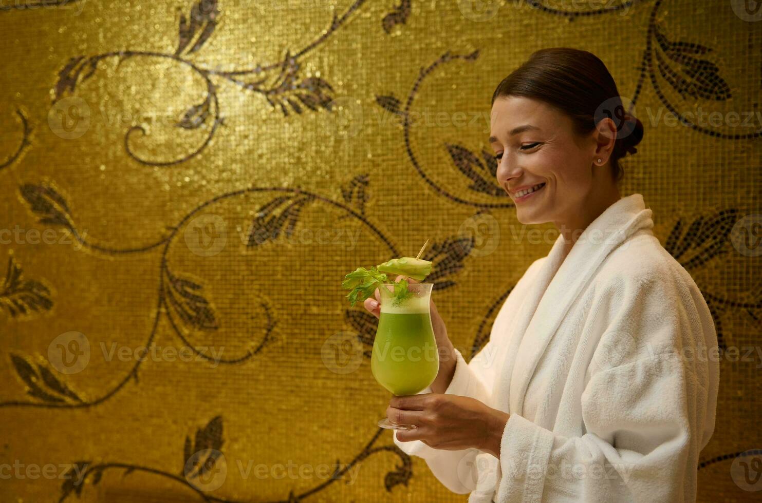 Smiling middle aged Caucasian pretty woman in white bathrobe, holding a glass with delicious healthy freshly squeezed vitamin juice in her hands, standing in a luxurious wellness center lounge area photo