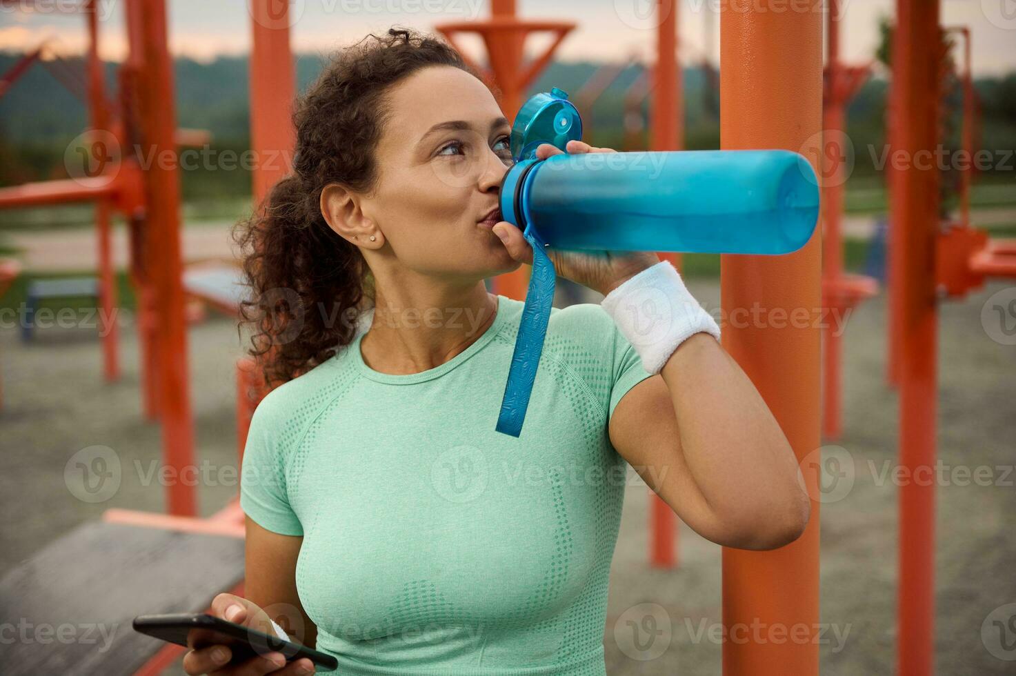 Attractive sporty Latin American woman with athletic physique holding a smartphone in one hand and drinking drinking water from a bottle after morning physical activities. Workout and fitness concept photo