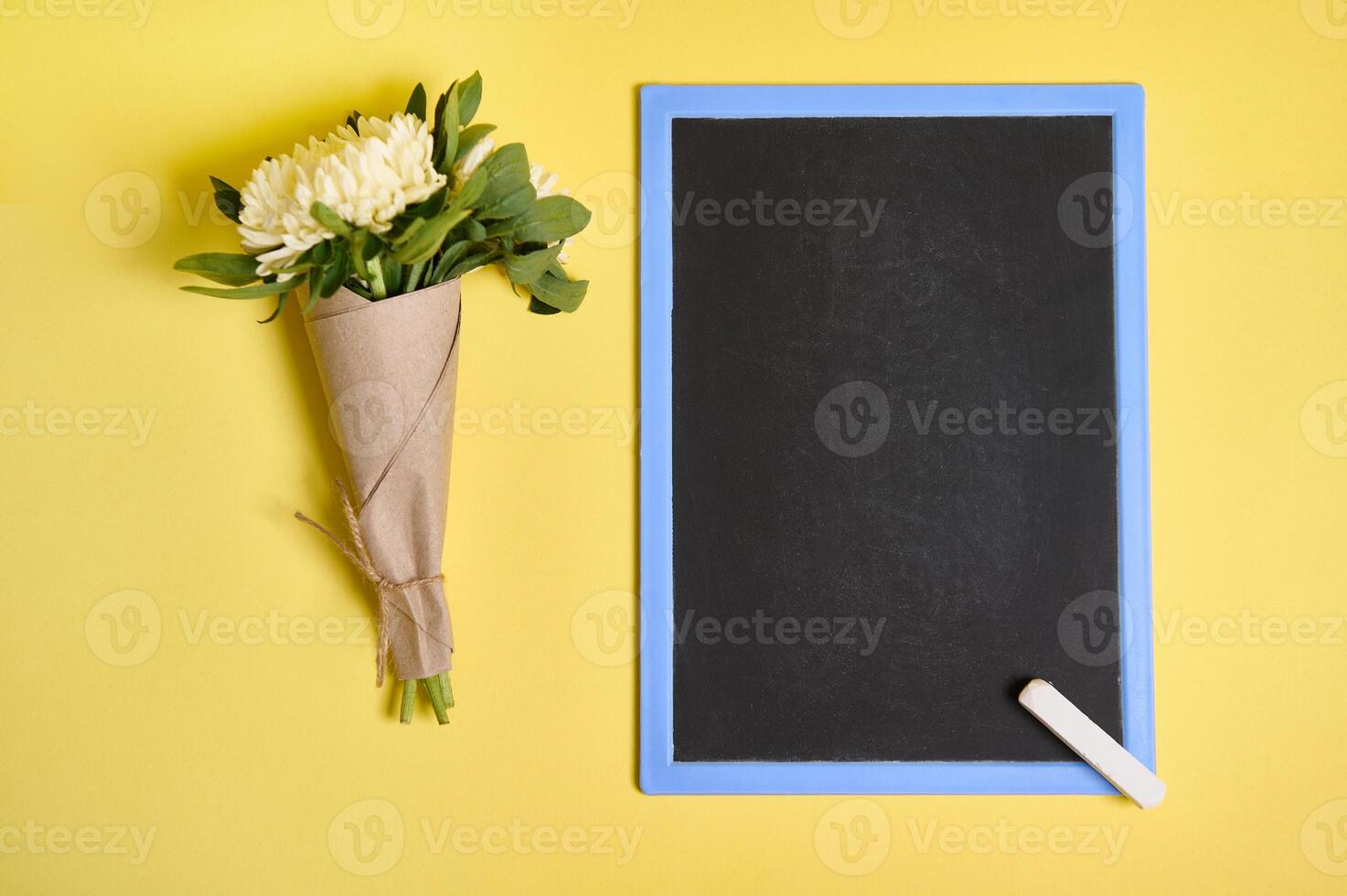 Flat lay composition of a delicate bouquet of autumn asters flowers in craft wrapping paper tied with a rope and blank empty chalkboard with space for text isolated on yellow background photo