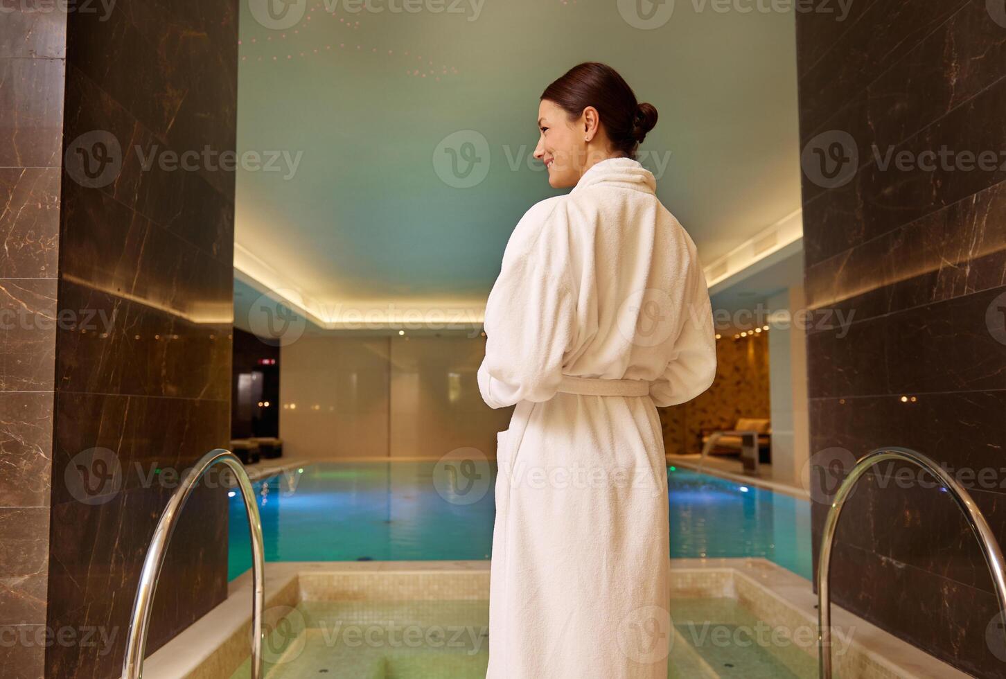 Rear view of a dark-haired beautiful woman in white terry robe standing by the pool and jacuzzi in luxury health spa enjoying relaxation and self-care. Beauty treatment, relaxation, body care concept photo