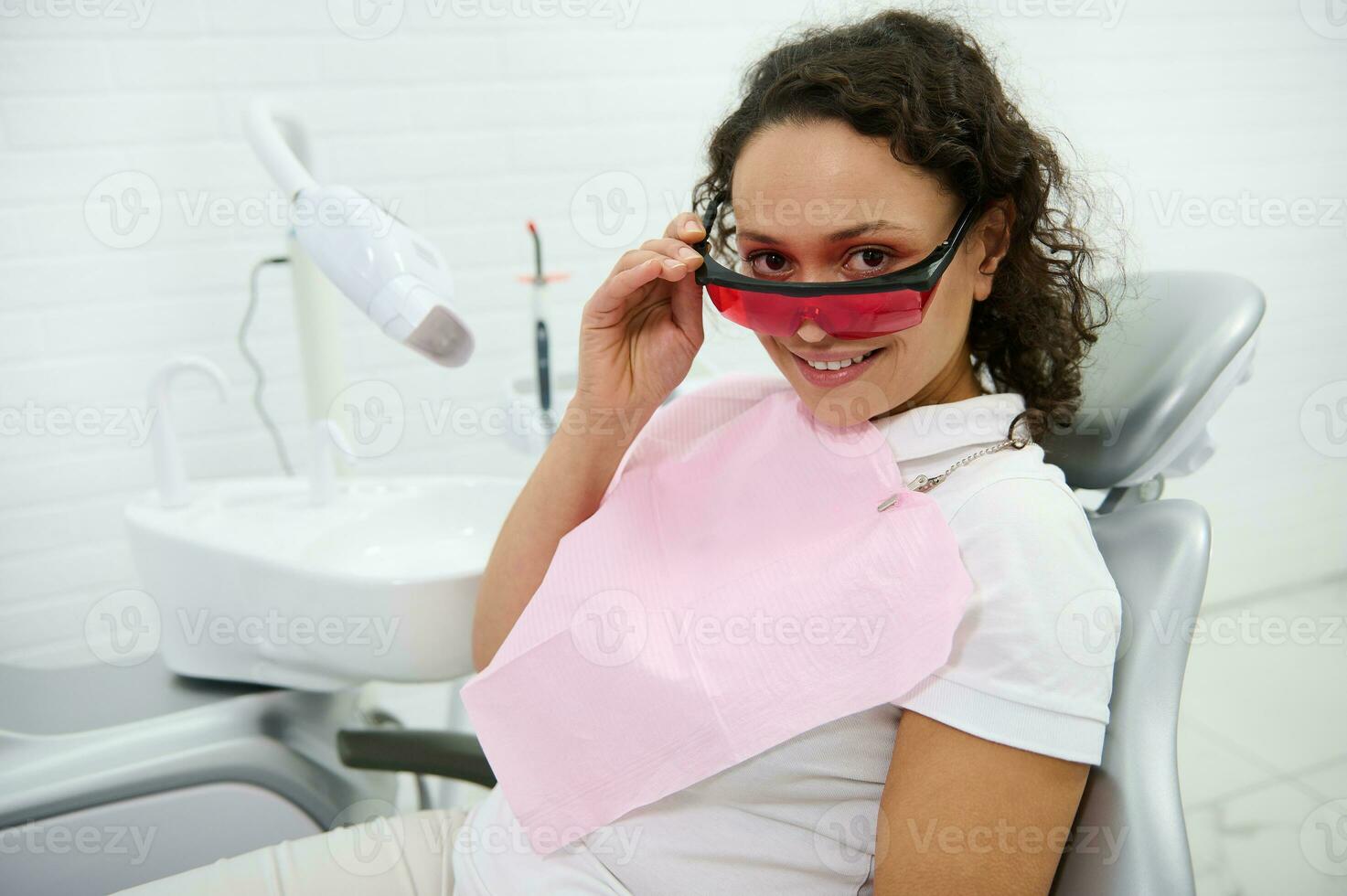 Charming woman wearing UV protective goggles smiles with beautiful toothy smile sitting in dentists chair, looking at camera after teeth whitening procedure with special UV light lamp in dental clinic photo