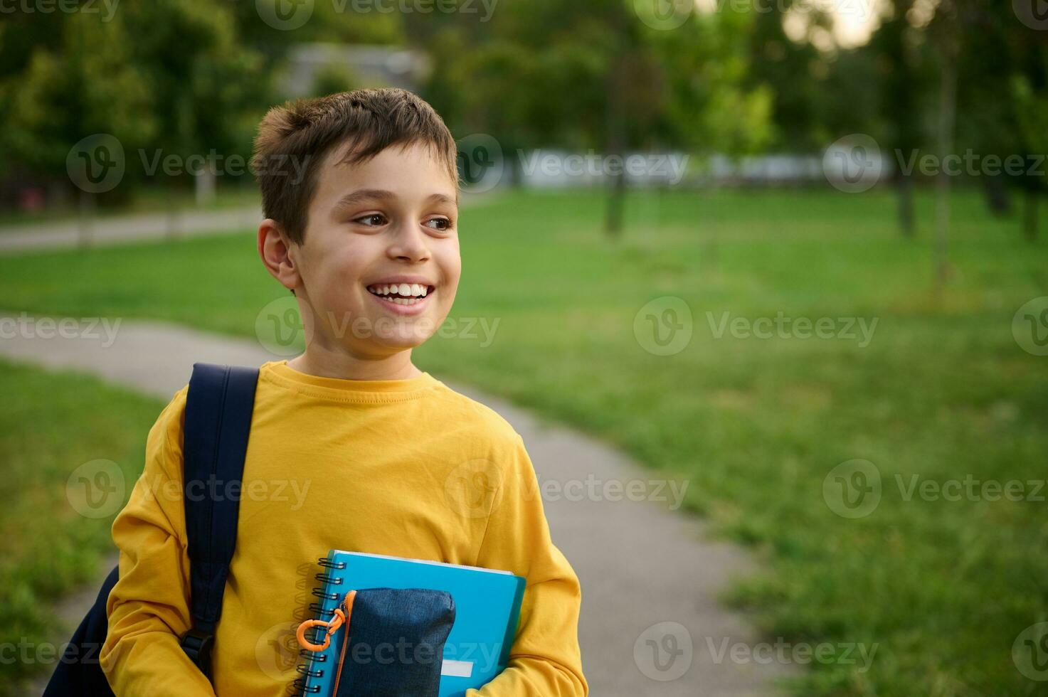 Portrait of handsome preadolescent schoolboy with backpack and pencil case with workbooks in his hands, looking away and smiling with toothy smile, enjoying the recreation after first day at school photo