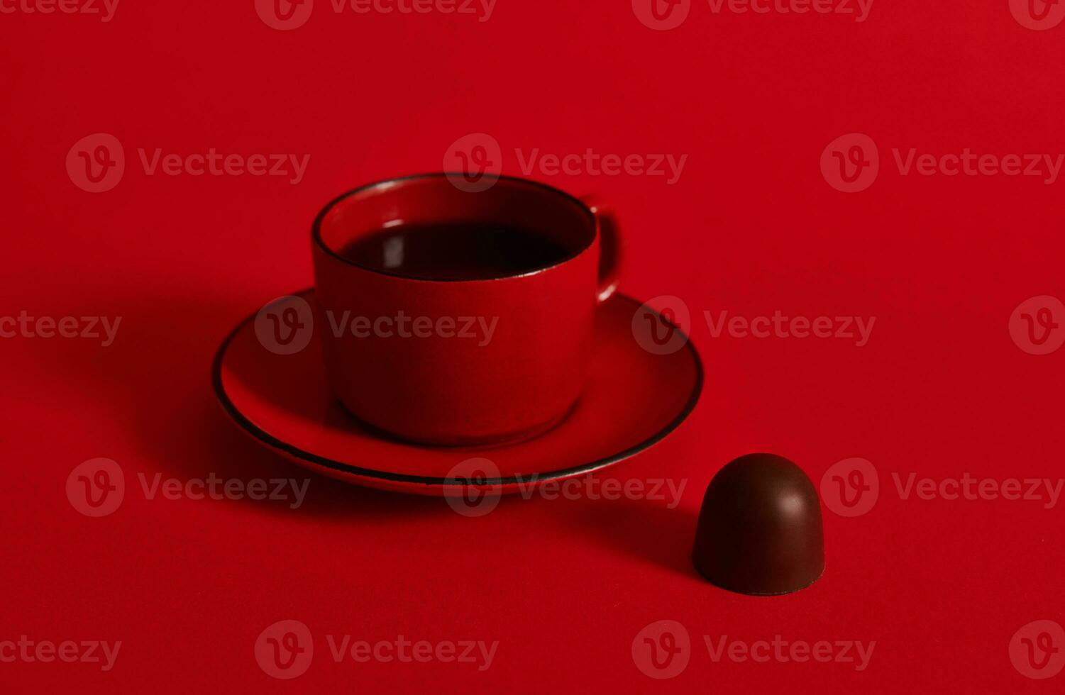 Closeup of a red ceramic cup of coffee on a saucer and a chocolate praline on red background with copy space . Concept of chocolate day photo