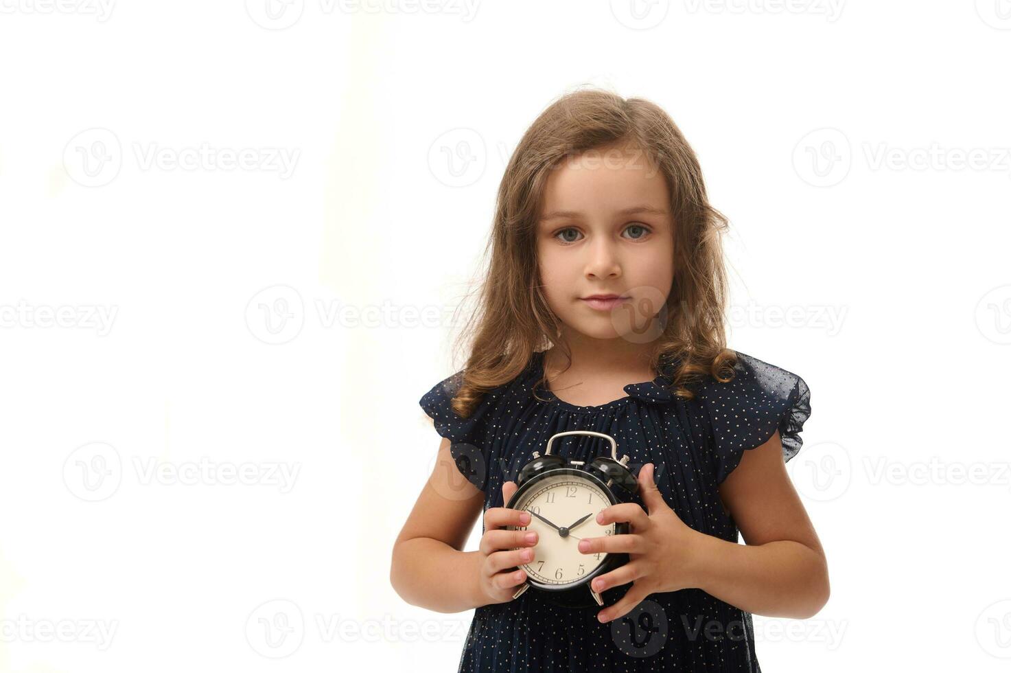 Adorable pretty 4 years old baby girl dressed in stylish dark blue evening attire, poses looking at the camera with an alarm clock in her hands, isolated against white background with copy space photo