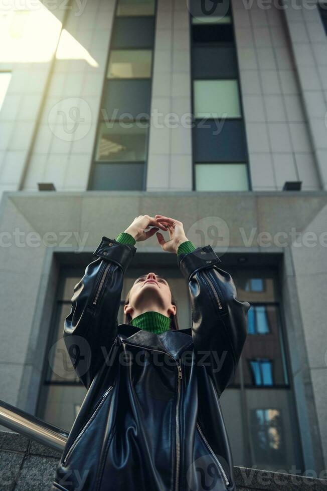 View from the bottom of fashionable young woman with stylish haircut holding smartphone in her raised hands against modern high-rise building. Connection, communication and business lifestyle concept photo