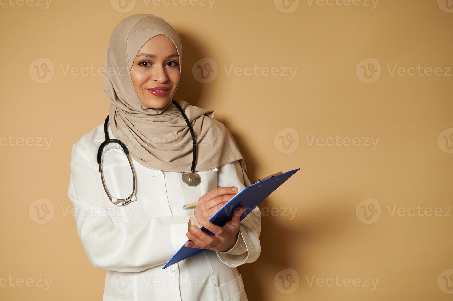 Female Muslim doctor wearing beige hijab cute smiles to camera and holds a pen and clipboard in hands. photo