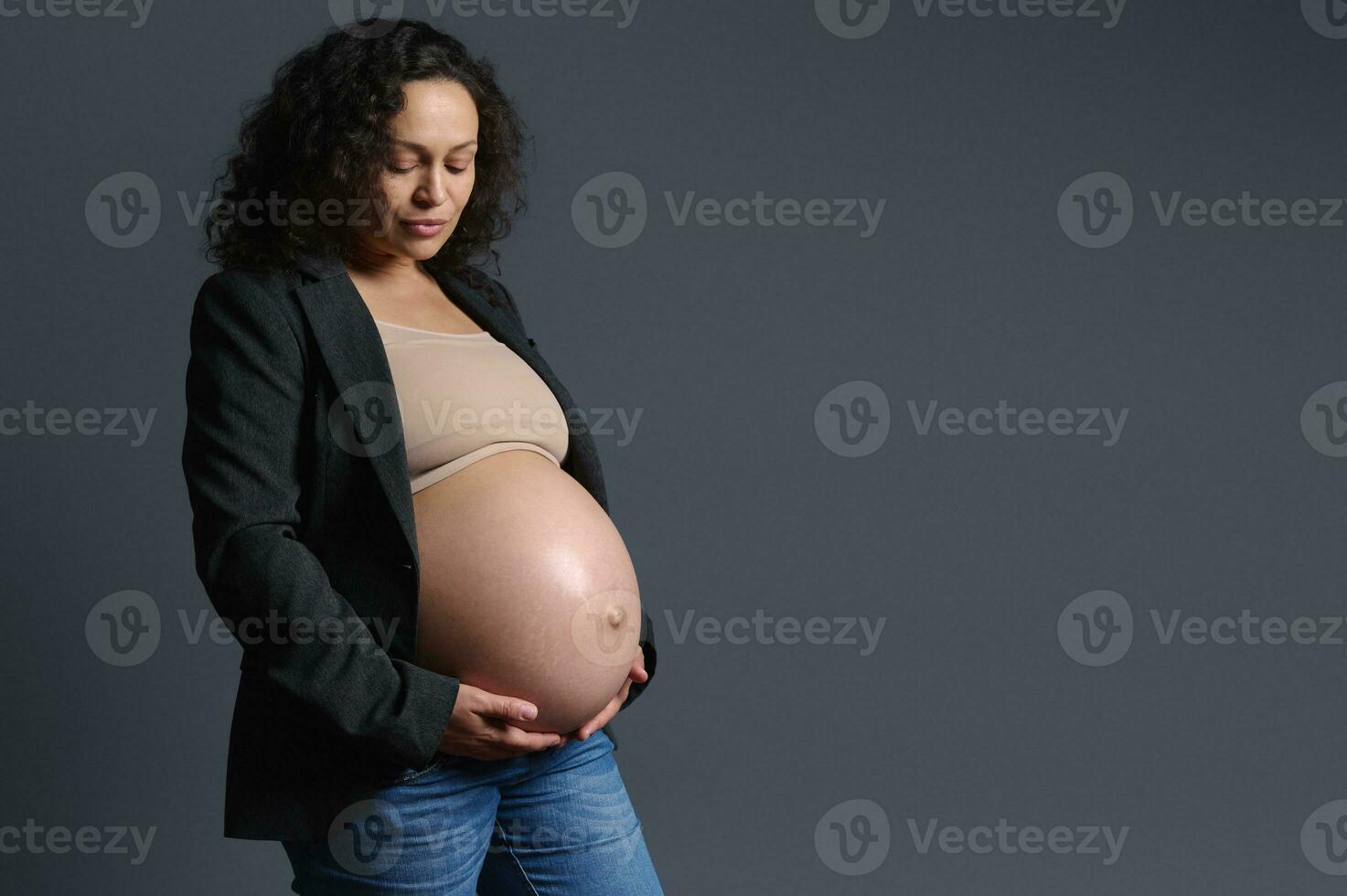 hermosa multiétnico embarazada mujer suavemente participación su desnudo barriga, aislado en gris antecedentes. el embarazo 3 trimestre foto