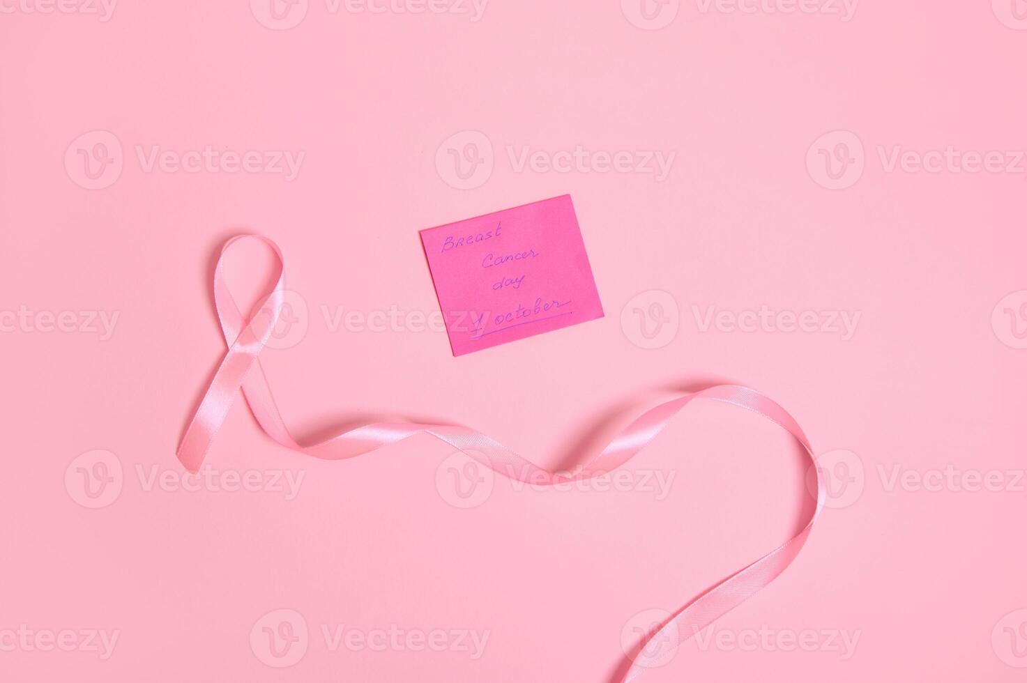 High angle view of a long pink ribbon with endless one end, and a paper note with lettering 1 October Breast Cancer Awareness , isolated on pink background with copy space. Flat lay photo