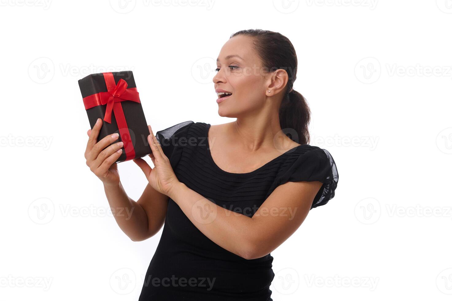 Beautiful happy Latin American woman dressed in black outfit poses against a white background , smiles toothy smile looking at a black gift box in her hands. Black Friday concept copy space for ad photo