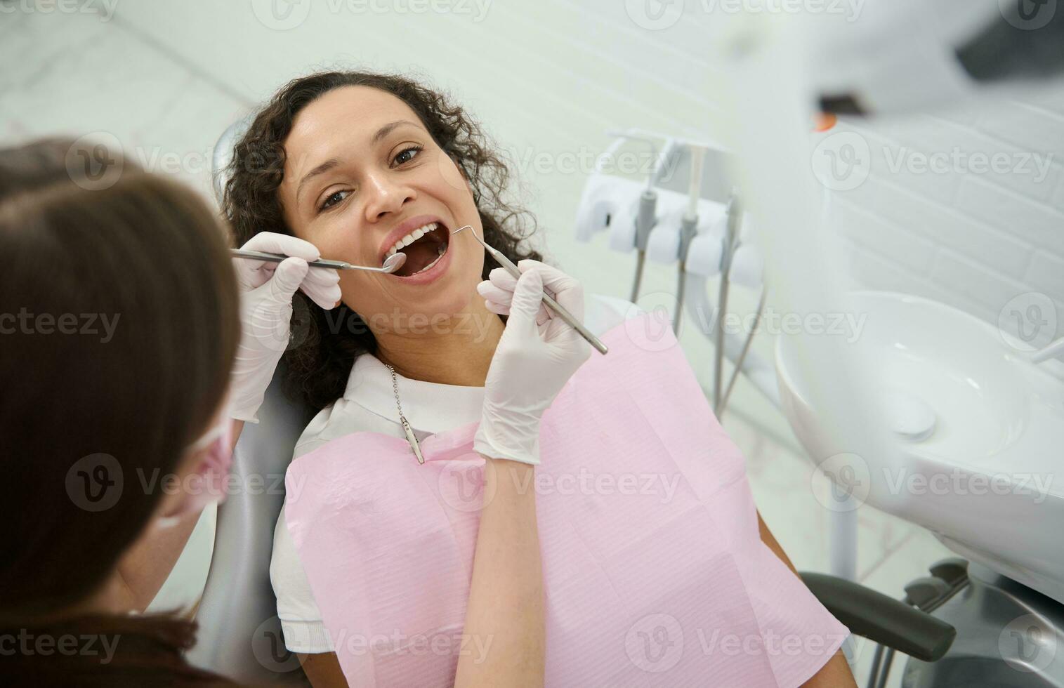 atractivo joven mujer con hermosa sonrisa se sienta en el del dentista silla con su boca abierto mientras el higienista examina o trata su dientes utilizando inoxidable acero dental instrumentos - espejo y Investigacion foto
