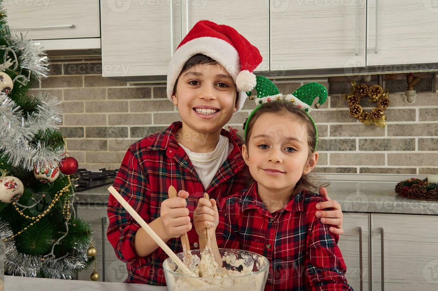 hermoso caucásico chico en Papa Noel claus sombrero sonrisas con alegre con dientes sonrisa abrazando su adorable pequeño hermana y mirando a cámara mientras amasadura y preparando masa para Navidad tradicional pastel foto