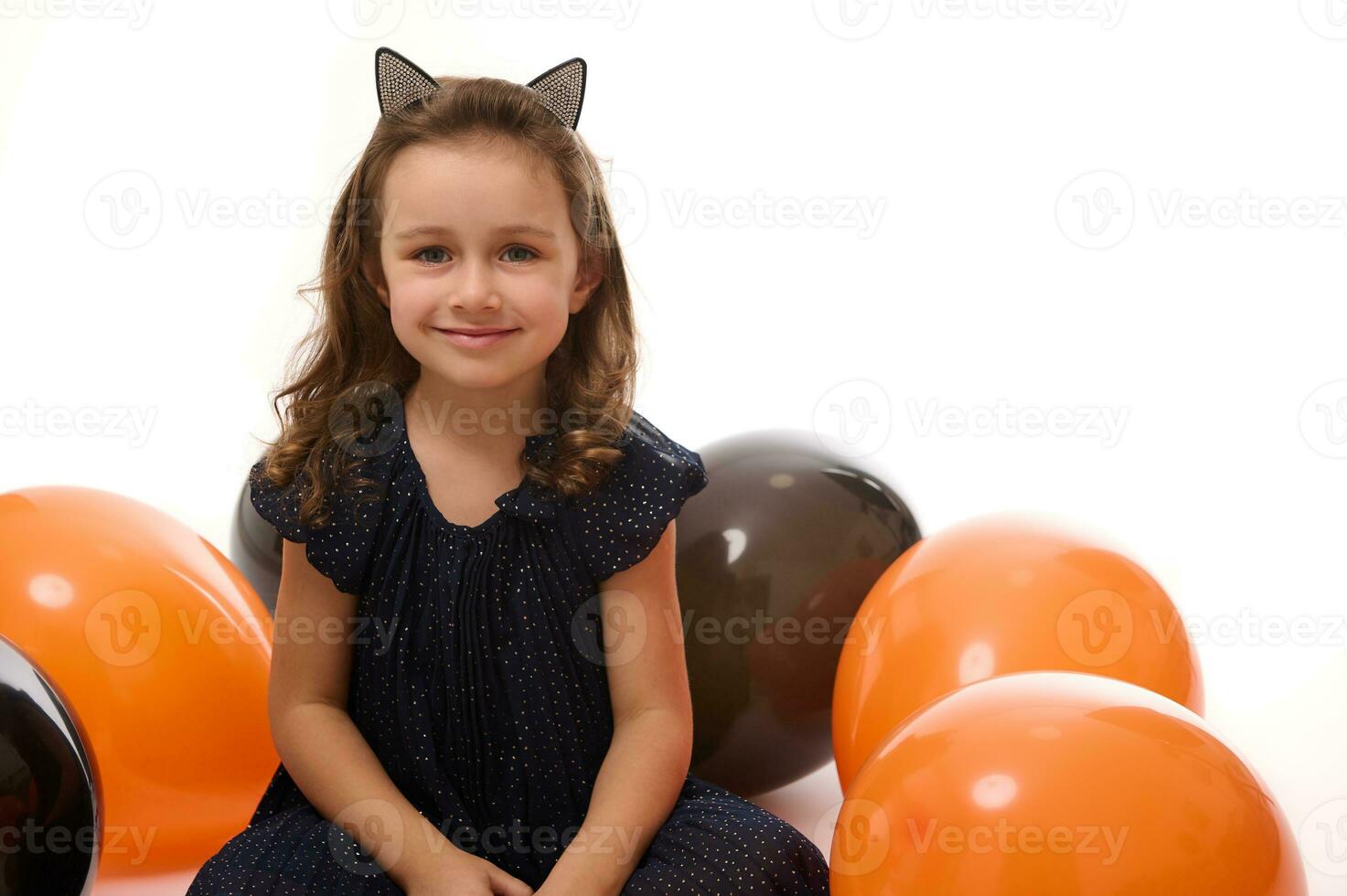 alegre atractivo adorable pequeño niña en negro carnaval vestir y aro con gato orejas sentado en un blanco superficie con bi de colores globos, sonrisas mirando a cámara. Víspera de Todos los Santos decoración. Copiar espacio foto