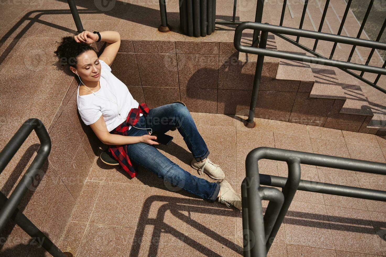 atractivo joven Hispano mujer escuchando a música en auriculares y teniendo descanso propensión en pasos de irreconocible edificio, disfrutando calentar soleado día foto