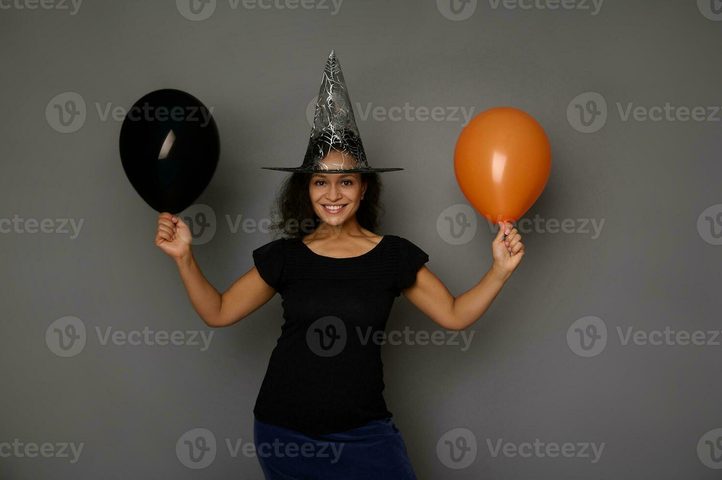 hermosa atractivo mujer vestido en bruja negro Víspera de Todos los Santos disfraz y mago sombrero, soportes en contra un gris antecedentes con naranja y negro aire globo, sonrisas mirando a cámara foto