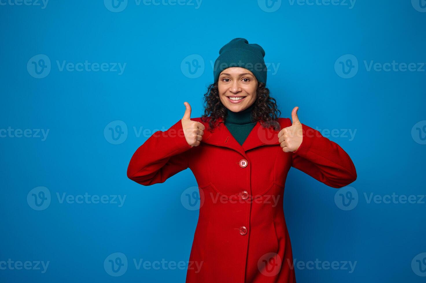 encantador maravilloso atractivo bonito mujer en brillante rojo Saco y verde sombrero sonriente con alegre con dientes sonrisa, mirando a cámara y demostración pulgares arriba, posando en contra azul antecedentes con Copiar espacio foto