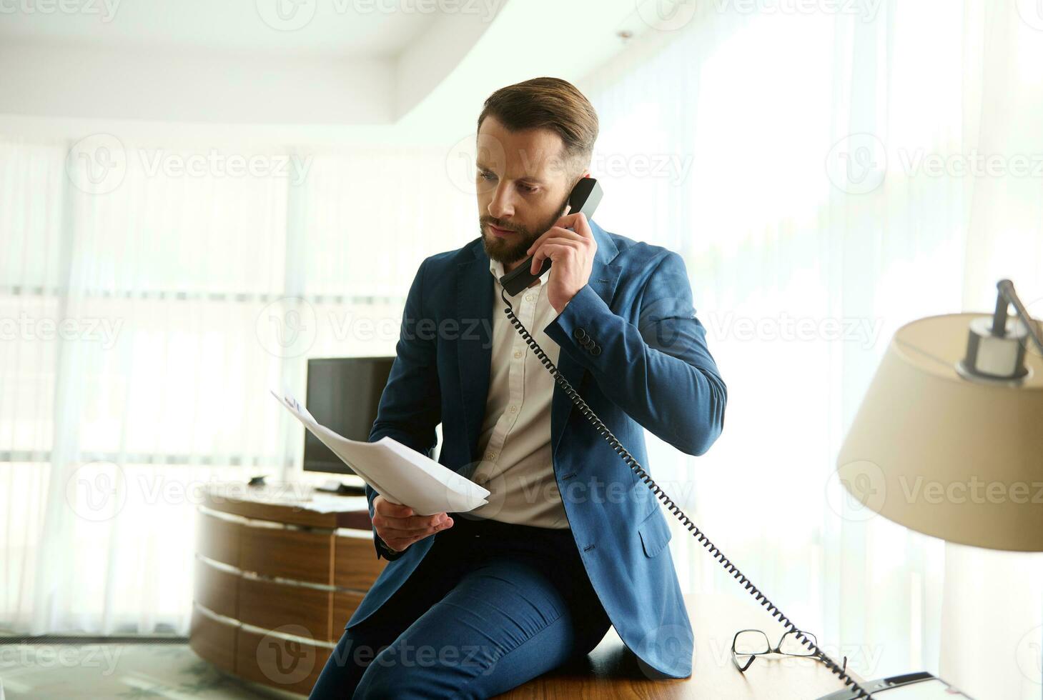 Thoughtful puzzled young business investor, businessman, finance director sitting on hotel table corner with documents and contract i hands and talking seriously on landline phone during business trip photo