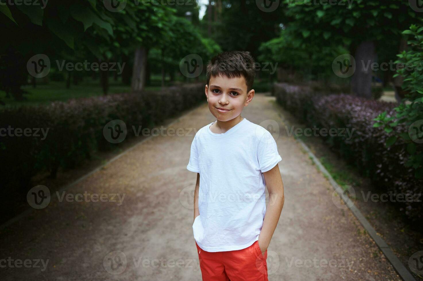 Portrait handsome Caucasian school age boy in white t-shirt, smiling looking at camera, standing in the alley of a park photo