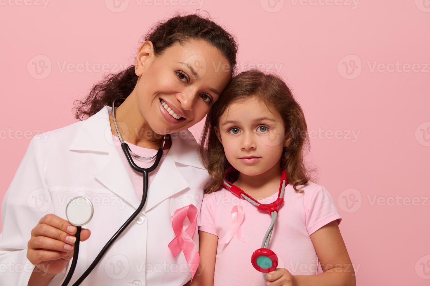 sonriente mezclado carrera médico y pequeño chica, ambos con pecho cáncer conciencia rosado cinta y fonendoscopio alrededor su cuello, demostración estetoscopio a cámara, aislado en rosado fondo, Copiar espacio. foto