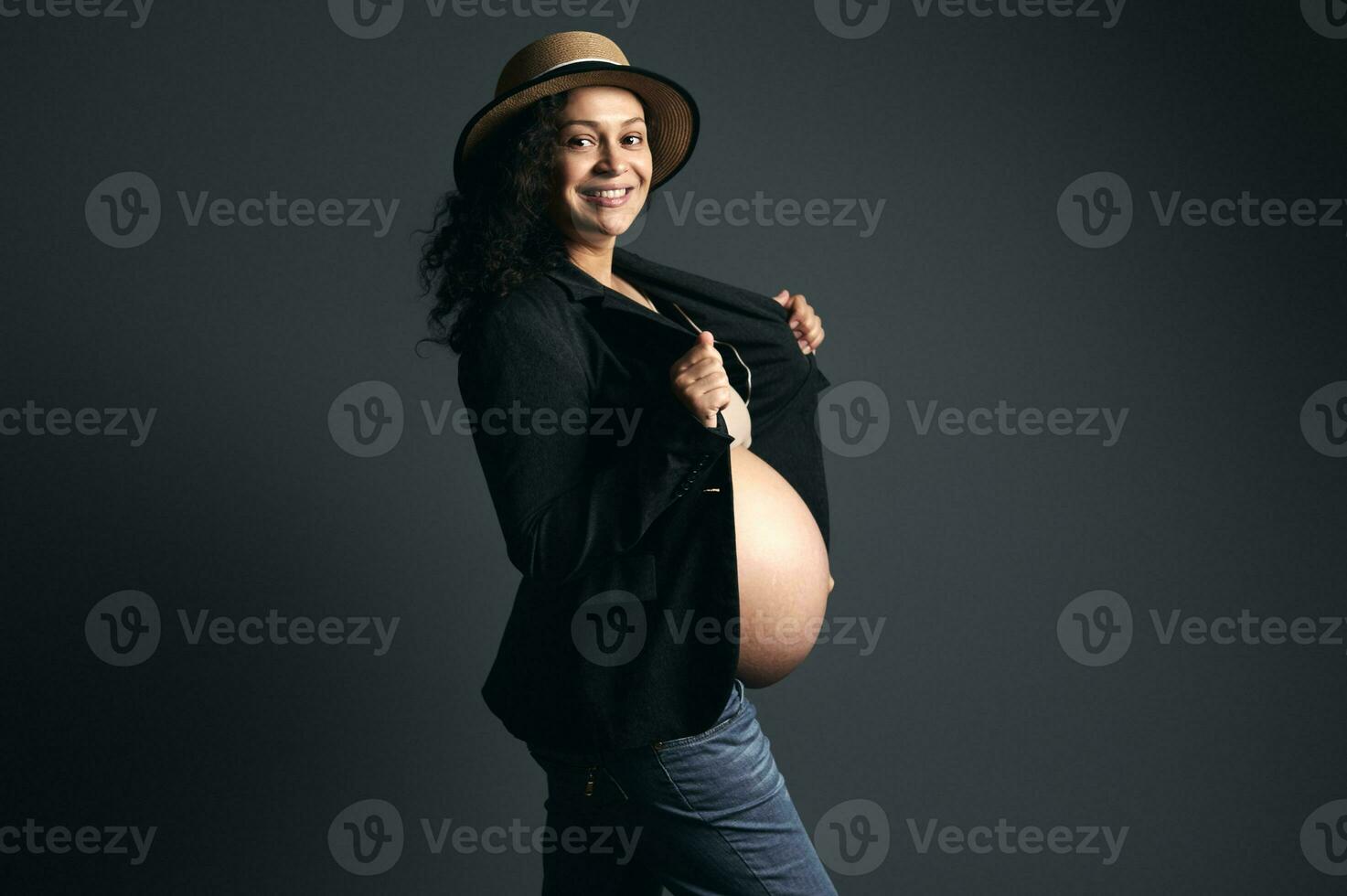 latín americano embarazada bonito mujer en Paja sombrero, elegante chaqueta de sport, sonrisas a cámara, posando desnudo barriga, estudio fondo foto