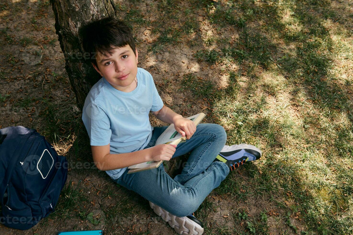 View from above of a school kid, adorable preteen boy in casual denim, reading book after school in the backyard photo