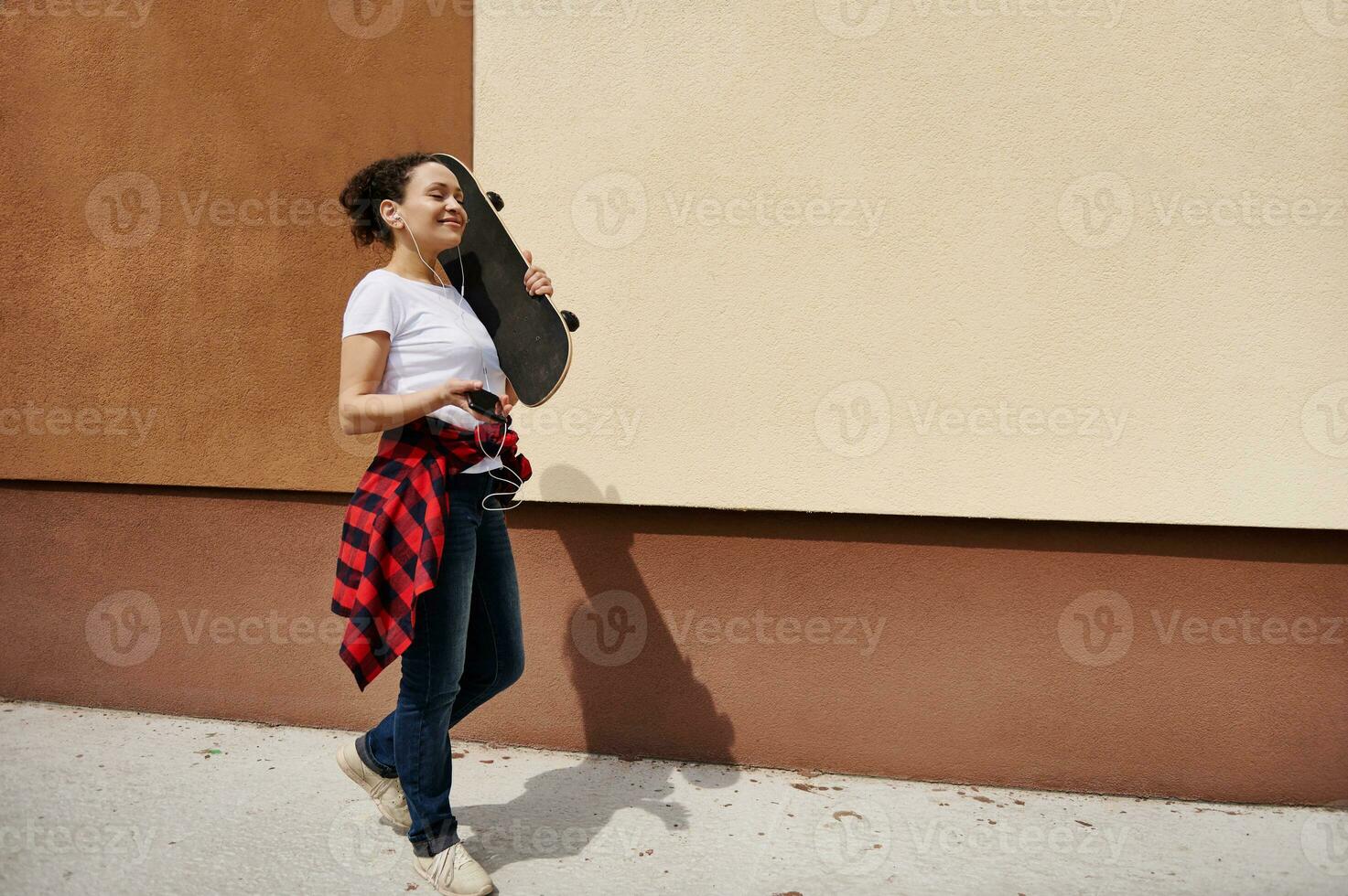 Happy woman holding wooden skateboard on shoulder walking down the street listening to music against colored wall background photo