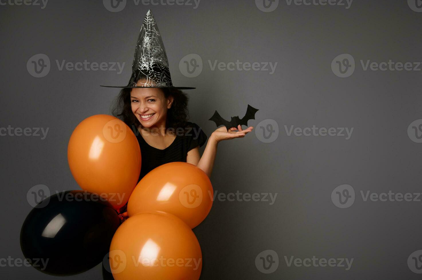 Beautiful mysterious smiling woman in wizard hat, dressed in black, holds orange black air balloons and handmade felt-cut black bat, looks at camera, poses against gray background, copy space . photo
