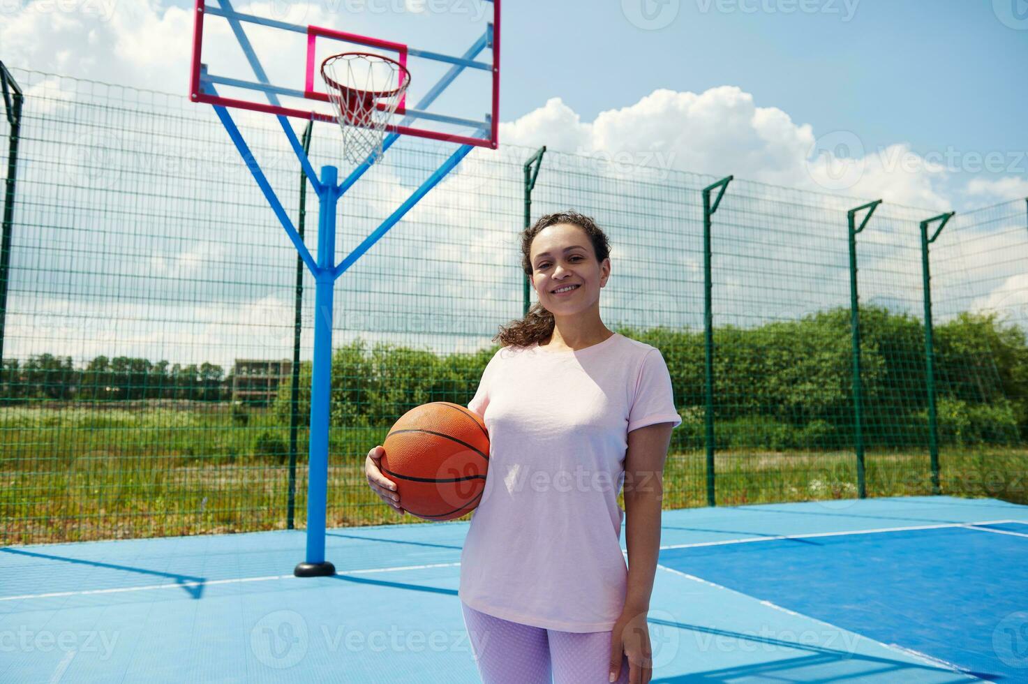 africano americano hembra cesta pelota jugador en pie en un Corte y sonrisas participación un baloncesto debajo brazos foto