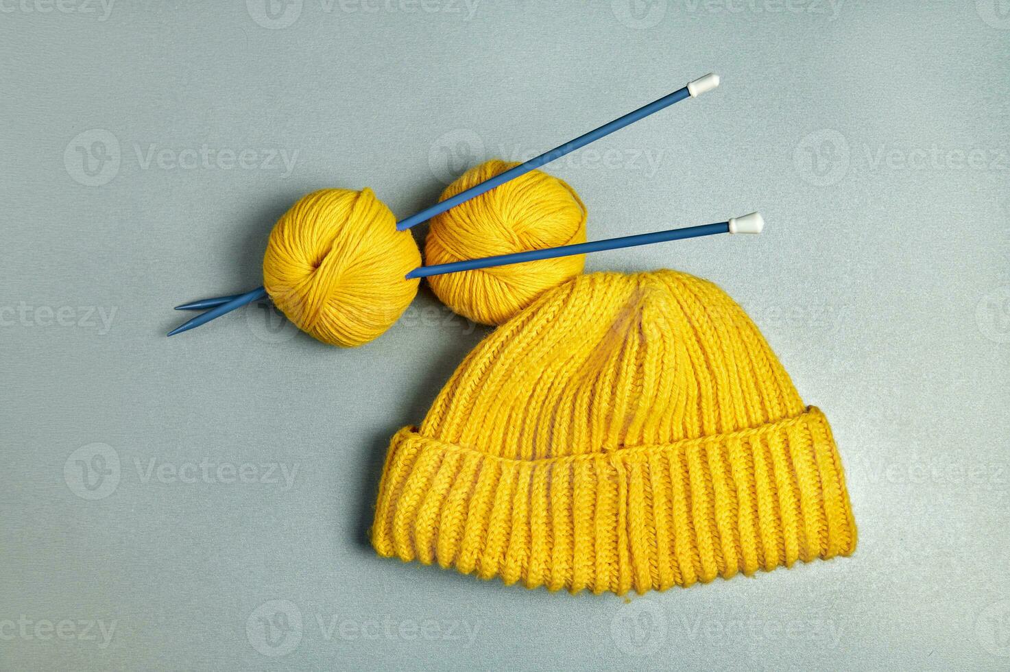 An item photograph of a yellow knitted woollen hat and two winding wool threads strung on long blue knitting needles on a grey background photo