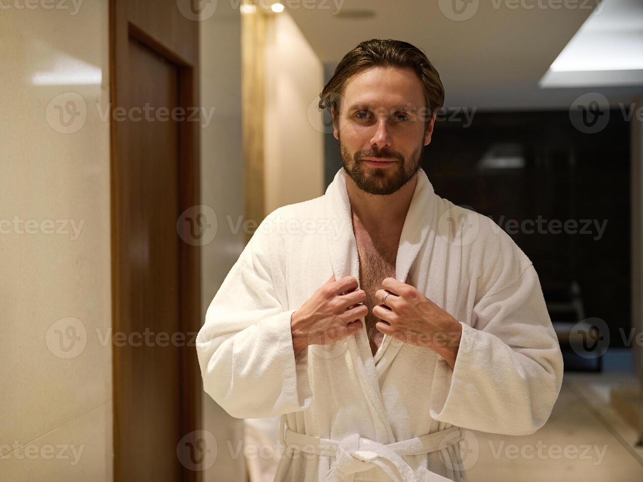 Attractive middle aged man with a charming look and a slight unshaven, handsome macho wearing white terry bathrobe, standing in a lobby of a luxury wellness spa complex, looking at camera photo