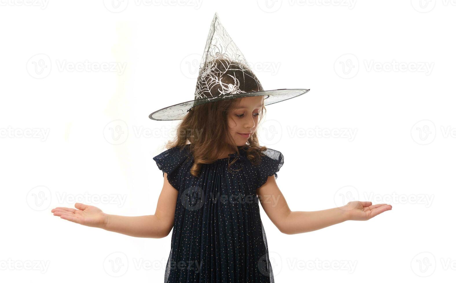 Portrait of pretty little witch girl wearing wizard hat, dressed in stylish carnival dress, gesturing, looking down at a copy space imaginary on the palm to insert an advertising. Halloween concept photo