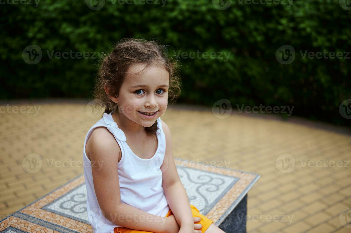 Caucasian adorable little child girl 4 years smiling looking at camera, sitting on a mosaic bench in the city park photo